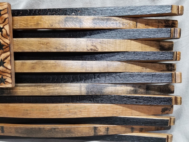 K&ouml;nnte beinhalten: Eine amerikanische Holzflagge aus wiederverwendeten Fassdauben. Die Flagge ist schwarz und braun mit einer Holzmaserung. Die Sterne sind in das Holz gebrannt.