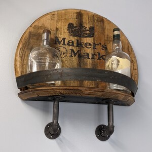 Op de afbeelding: Een houten plank met een Maker's Mark logo, twee doorzichtige glazen flessen en zwarte metalen pijpfittingen. De plank is gemaakt van gerecycled hout en heeft een rustieke uitstraling.