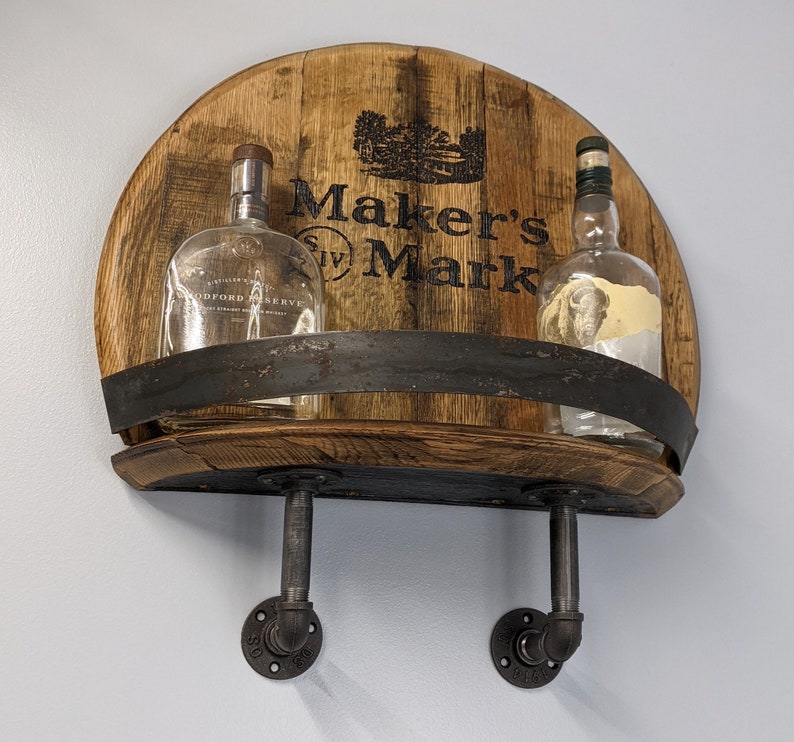 Op de afbeelding: Een houten plank met een Maker's Mark logo, twee doorzichtige glazen flessen en zwarte metalen pijpfittingen. De plank is gemaakt van gerecycled hout en heeft een rustieke uitstraling.