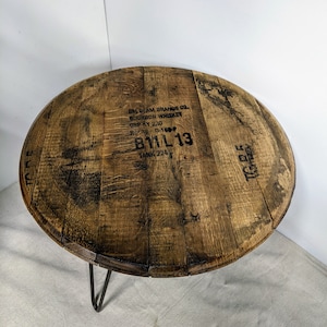 Pode incluir: Uma mesa redonda constru&iacute;da a partir de uma tampa de barril de bourbon de madeira recuperada com pernas de metal escuro em forma de gancho. O tampo apresenta veios de madeira natural e texto preto estampado que diz JIM BEAM BRANDS CO. BOURBON.