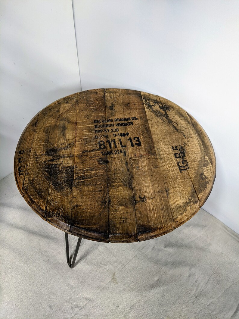 Pode incluir: Uma mesa redonda constru&iacute;da a partir de uma tampa de barril de bourbon de madeira recuperada com pernas de metal escuro em forma de gancho. O tampo apresenta veios de madeira natural e texto preto estampado que diz JIM BEAM BRANDS CO. BOURBON.