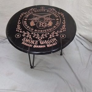Pode incluir: Uma mesa redonda preta feita de uma tampa de barril com tr&ecirc;s pernas de metal finas. O topo apresenta um gr&aacute;fico cor de cobre de dois rev&oacute;lveres cruzados, estrelas, vinhas florais e o texto Smoke Wagon Bourbon, Bibamus Moriendum Est e Straight Bourbon Whiskey.
