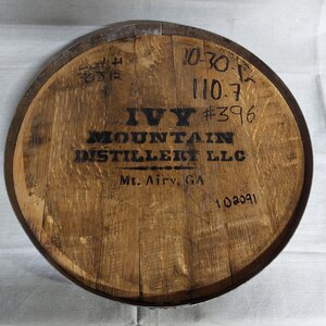 May include: A wooden barrel head with a black logo that reads "Ivy Mountain Distillery LLC" and "Mt. Airy, GA". The barrel head also has several handwritten numbers, including "10-30-12", "110.7", "#396", and "102091".