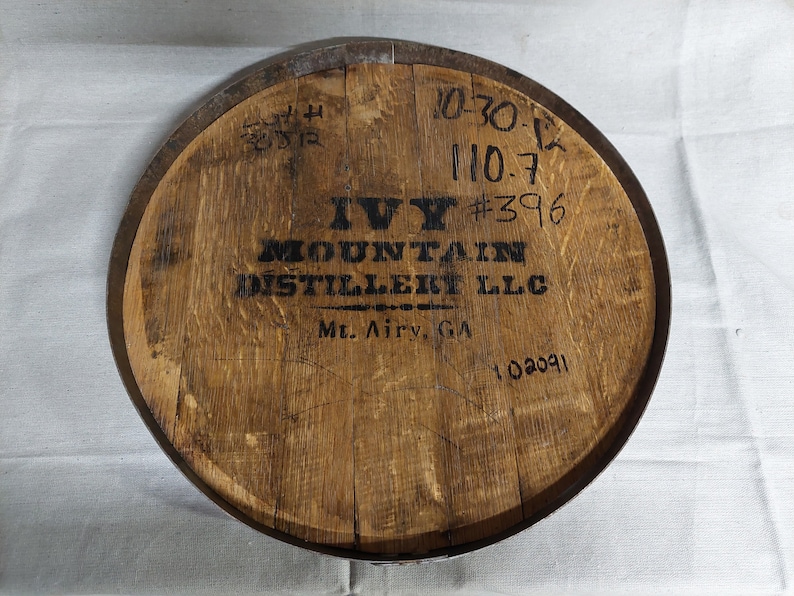 May include: A wooden barrel head with a black logo that reads "Ivy Mountain Distillery LLC" and "Mt. Airy, GA". The barrel head also has several handwritten numbers, including "10-30-12", "110.7", "#396", and "102091".