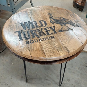 Pode incluir: Uma mesa lateral redonda constru&iacute;da com aduelas de barril de madeira reciclada com um acabamento natural castanho claro. O tampo apresenta um gr&aacute;fico impresso a preto de um peru e o texto WILD TURKEY BOURBON. Apoia-se em tr&ecirc;s pernas de metal preto.