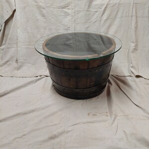 May include: A circular coffee table crafted from a reclaimed wooden barrel. The barrel is dark brown with metal bands and a clear glass top. The table is a unique piece of furniture.