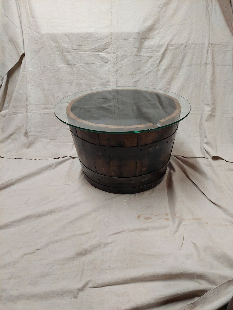 May include: A circular coffee table crafted from a reclaimed wooden barrel. The barrel is dark brown with metal bands and a clear glass top. The table is a unique piece of furniture.
