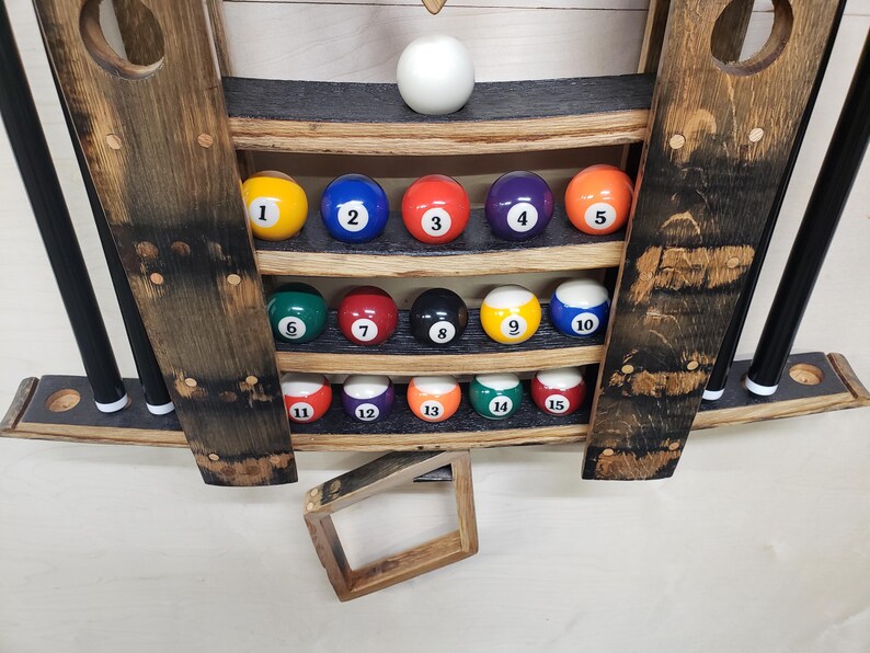 May include: A wooden wall-mounted rack for storing billiard balls and cues. The rack has four shelves with 15 billiard balls in place, numbered 1 through 15. The rack is made of dark wood with a black finish. Two cues are stored in the rack.