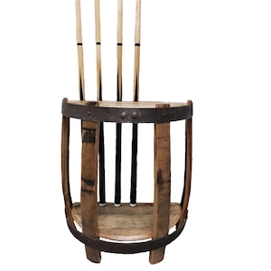 May include: A rustic wooden pool cue rack made from a repurposed barrel. The rack holds four cues, with a natural wood finish and metal bands. The cues are light-colored with black accents.