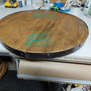 May include: A round wooden tabletop with a metal band around the edge. The wood is a light brown colour and the metal band is a dark grey colour. The tabletop has a green label with text that reads "ALL WOOD" and "11.5 GALLON".