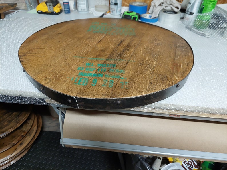 May include: A round wooden tabletop with a metal band around the edge. The wood is a light brown colour and the metal band is a dark grey colour. The tabletop has a green label with text that reads "ALL WOOD" and "11.5 GALLON".