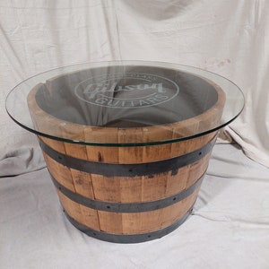 May include: A circular coffee table constructed from a reclaimed wooden barrel. The barrel is secured with metal bands and features a clear glass top. The glass is etched with the words "Gibson Guitars".