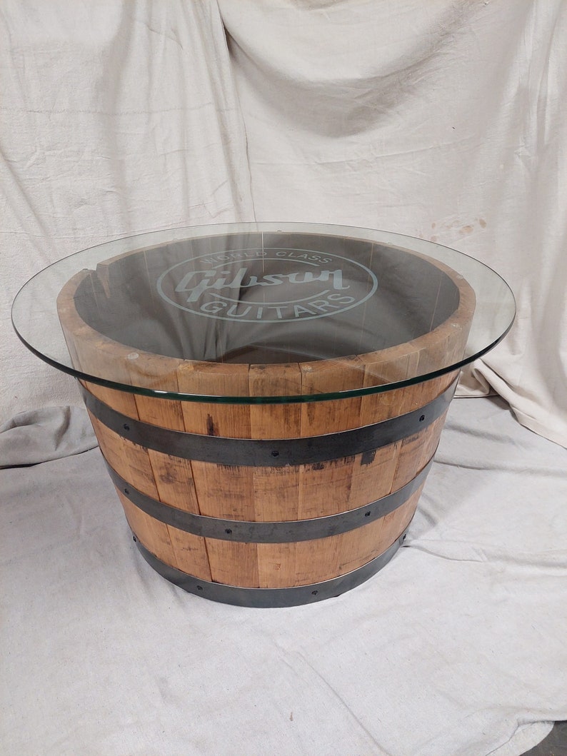 May include: A circular coffee table constructed from a reclaimed wooden barrel. The barrel is secured with metal bands and features a clear glass top. The glass is etched with the words "Gibson Guitars".