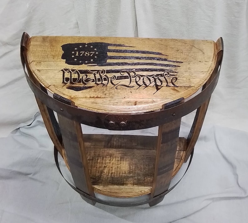 K&ouml;nnte beinhalten: Ein halbkreisf&ouml;rmiger Beistelltisch aus recycelten Holzfassdauben und Metallreifen. Die Tischplatte zeigt eine eingebrannte Grafik einer stilisierten amerikanischen Flagge mit der Jahreszahl 1787 und dem Schriftzug We The People.
