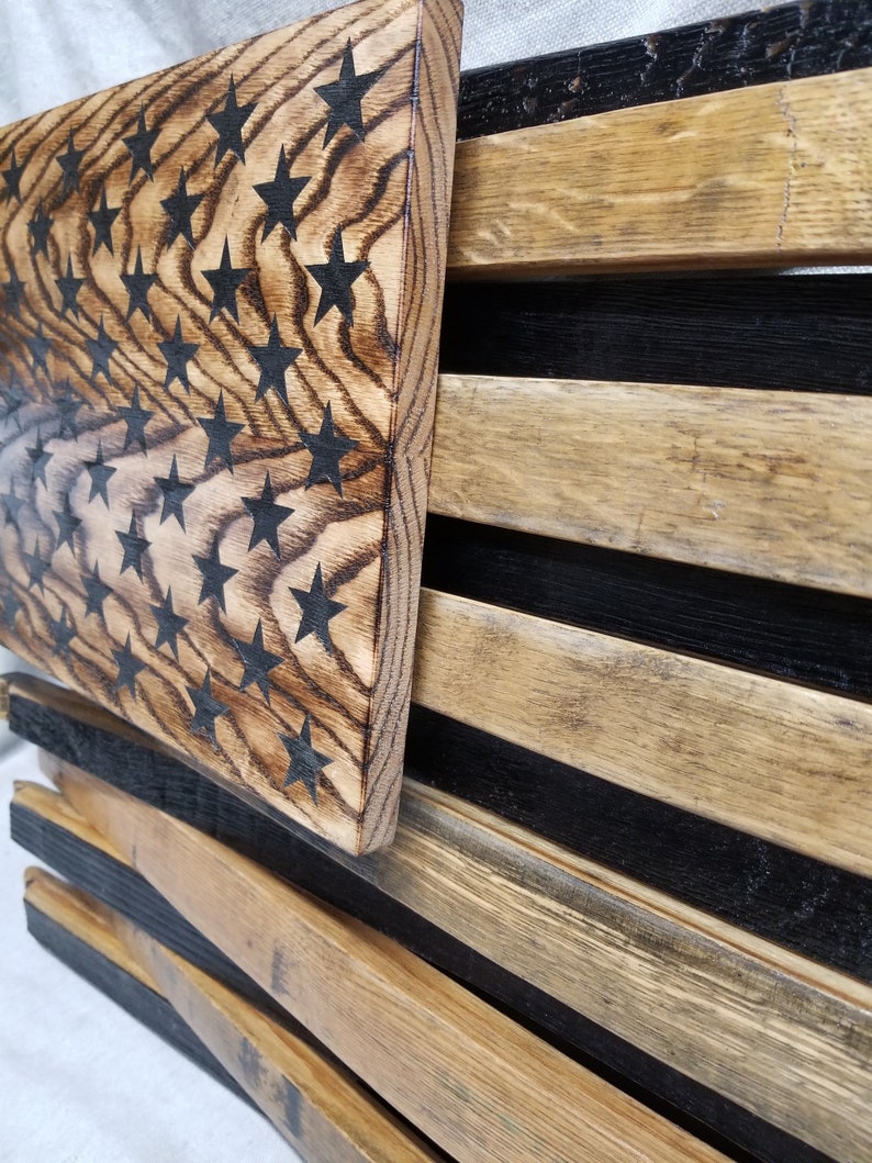 K&ouml;nnte beinhalten: Eine amerikanische Holzflagge aus recyceltem Holz mit schwarzen Sternen auf hellbraunem Hintergrund. Die Flagge wird an einer Wand gezeigt.