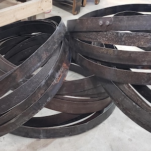 Pode incluir: Uma cole&ccedil;&atilde;o de aros de barril de metal escuro e desgastado, alguns com rebites vis&iacute;veis. Os aros est&atilde;o dispostos em pilhas soltas, sugerindo um tema r&uacute;stico ou industrial. O metal tem uma apar&ecirc;ncia desgastada e envelhecida.