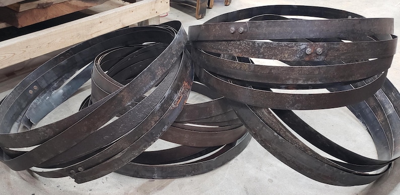 Pode incluir: Uma cole&ccedil;&atilde;o de aros de barril de metal escuro e desgastado, alguns com rebites vis&iacute;veis. Os aros est&atilde;o dispostos em pilhas soltas, sugerindo um tema r&uacute;stico ou industrial. O metal tem uma apar&ecirc;ncia desgastada e envelhecida.