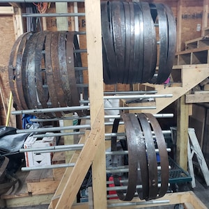 Pode incluir: Uma cole&ccedil;&atilde;o de aros de barril de metal escuro e desgastado, exibidos em um suporte de madeira. Os aros variam em tamanho e mostram sinais de idade e uso, sugerindo um tema r&uacute;stico ou industrial. O fundo inclui estruturas de madeira e uma escada.