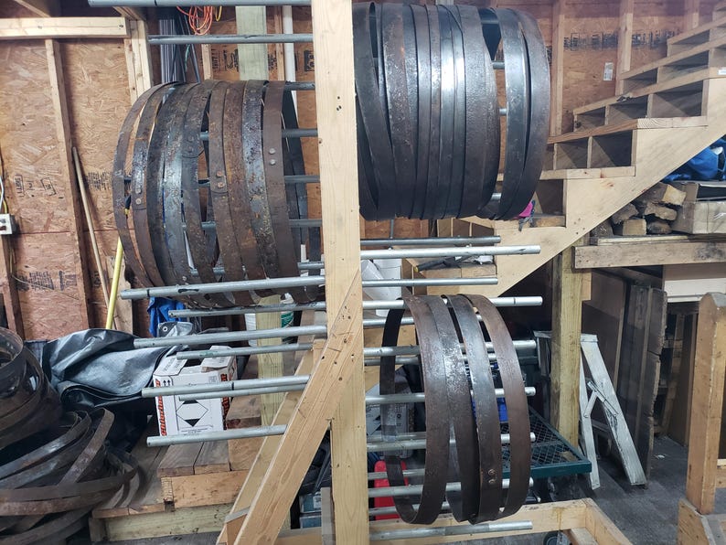 Pode incluir: Uma cole&ccedil;&atilde;o de aros de barril de metal escuro e desgastado, exibidos em um suporte de madeira. Os aros variam em tamanho e mostram sinais de idade e uso, sugerindo um tema r&uacute;stico ou industrial. O fundo inclui estruturas de madeira e uma escada.