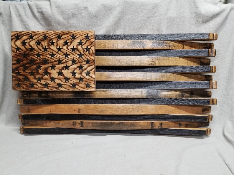 K&ouml;nnte beinhalten: Eine amerikanische Flagge aus Holz, hergestellt aus wiederverwendeten Bourbon-Fassdauben. Die Sterne sind in das Holz gebrannt und die Streifen wechseln sich in Dunkelbraun und Hellbraun ab.