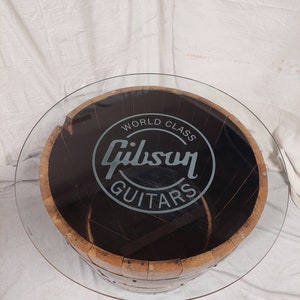 May include: Circular coffee table featuring a glass top and a wooden barrel base. The barrel is dark brown with metal bands. The text "World Class Gibson Guitars" is displayed on the table's surface.