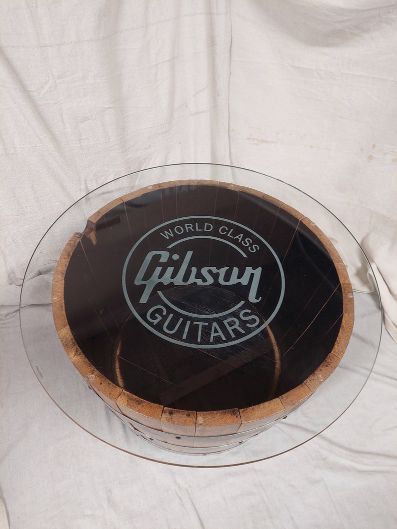 May include: Circular coffee table featuring a glass top and a wooden barrel base. The barrel is dark brown with metal bands. The text "World Class Gibson Guitars" is displayed on the table's surface.