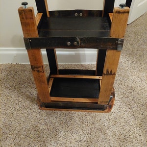 May include: A two-tiered wooden stand made from repurposed barrel staves. The stand has a black stained finish and features metal brackets and screws. The stand is approximately 61 cm tall.