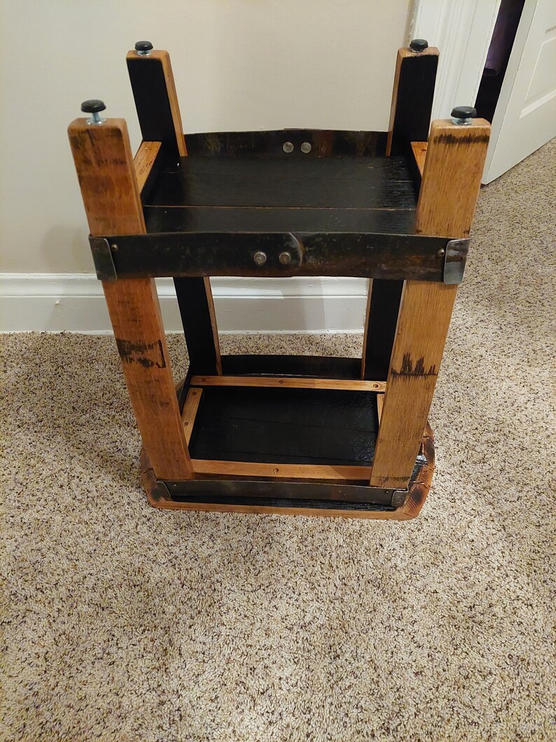 May include: A two-tiered wooden stand made from repurposed barrel staves. The stand has a black stained finish and features metal brackets and screws. The stand is approximately 61 cm tall.
