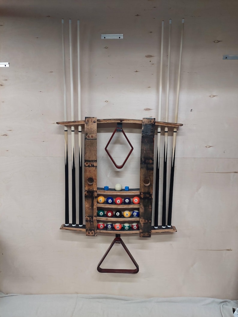 May include: A wooden wall-mounted pool cue rack with a rack for billiard balls. The rack is made from reclaimed wood barrels and features a red felt triangle rack. The rack holds 15 billiard balls and 5 pool cues.