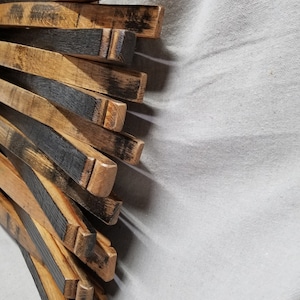 K&ouml;nnte beinhalten: Ein Stapel aus wiederverwendeten Holzfassdauben, mit nat&uuml;rlicher Holzfarbe und schwarzen Brandspuren. Die Dauben sind f&auml;cherf&ouml;rmig angeordnet, wobei die Enden der Dauben sichtbar sind.