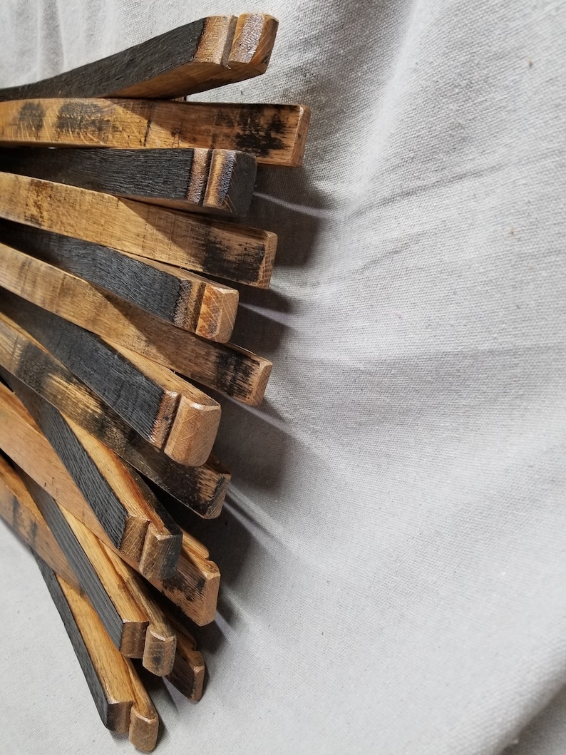 K&ouml;nnte beinhalten: Ein Stapel aus wiederverwendeten Holzfassdauben, mit nat&uuml;rlicher Holzfarbe und schwarzen Brandspuren. Die Dauben sind f&auml;cherf&ouml;rmig angeordnet, wobei die Enden der Dauben sichtbar sind.