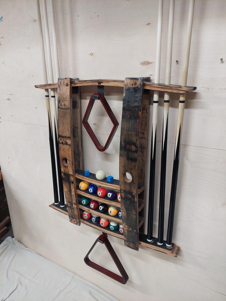 May include: A wooden wall-mounted pool cue rack with a rack for billiard balls. The rack is made from reclaimed wood and has a rustic look. It features a red felt triangle rack and a diamond-shaped rack for holding a cue stick.