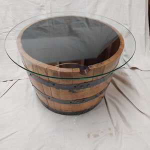 May include: A round coffee table with a glass top, set on a repurposed wooden barrel. The barrel is made of wooden staves held together by metal bands. The table is approximately 76 cm in diameter.