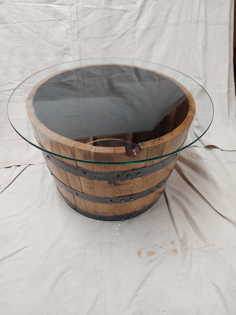 May include: A round coffee table with a glass top, set on a repurposed wooden barrel. The barrel is made of wooden staves held together by metal bands. The table is approximately 76 cm in diameter.