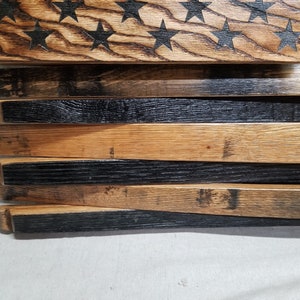 K&ouml;nnte beinhalten: Amerikanische Flagge aus wiederverwendeten Holzfassdauben. Die Flagge wird horizontal mit den Sternen oben und den Streifen unten angezeigt. Die Sterne sind in das Holz gebrannt und die Streifen sind schwarz gestrichen.