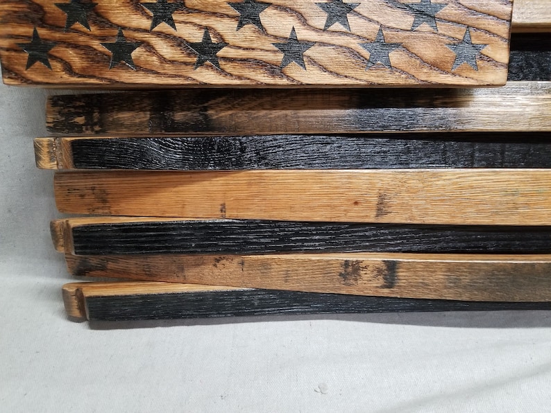 K&ouml;nnte beinhalten: Amerikanische Flagge aus wiederverwendeten Holzfassdauben. Die Flagge wird horizontal mit den Sternen oben und den Streifen unten angezeigt. Die Sterne sind in das Holz gebrannt und die Streifen sind schwarz gestrichen.