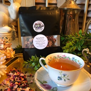 May include: A black bag of loose leaf tea called "Spring Sunrise" with a floral design. The tea is made in a home kitchen and weighs 1.55 oz (44g). The tea is displayed on a gold tray with a teacup and saucer, a teapot, and a white rabbit figurine.