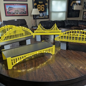 May include: A collection of yellow bridge models with black roadbeds and small white lights. The bridges are displayed on a dark wooden table. The bridges are supported by gray rectangular blocks.