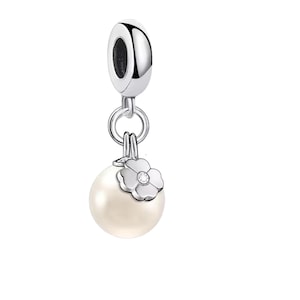 Puede incluir: Un charm de tono plateado con un núcleo de metal pulido y un colgante. El colgante consiste en una esfera blanca lisa similar a una perla, rematada con un pequeño adorno de flor de plata de cuatro pétalos que contiene una diminuta piedra clara.