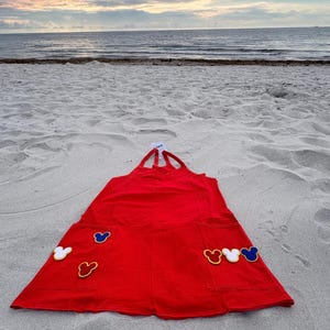 May include: A bright red apron featuring a halter-style neck strap and two large front pockets. Each pocket is decorated with three small, embroidered mouse-ear silhouettes in yellow, white, and blue, creating a playful and colorful design.