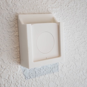 以下が含まれることがあります： 円形の中心ボタンが付いた、白い長方形の壁掛けデバイス。デバイスはフレーム内にセットされ、テクスチャ加工された白い壁に取り付けられています。デバイスの下部に小さな黒い点があります。