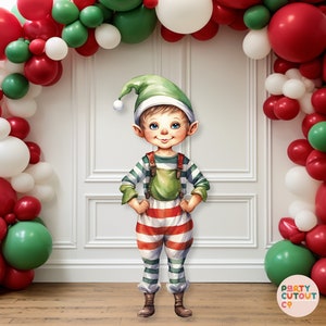 May include: A festive Christmas scene featuring a cartoon elf cutout standing in front of a white wall decorated with a red, white and green balloon arch. The elf is wearing a green hat, a red and white striped shirt and green trousers. The balloon arch is made of red, white and green balloons.