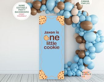 May include: A light blue party backboard with the text "Jaxon is one little cookie" and cookie illustrations. The backboard is decorated with cookie-themed accents and is part of a party cutout set. A balloon arch in blue, brown, and cream colors is in the background.