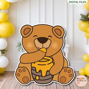 May include: A brown bear cartoon character holding a honey pot with a yellow lid and dripping honey. The bear is smiling and has a happy expression. The background is a white wall with yellow and white balloons.