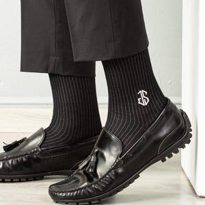May include: A pair of black dress socks with a white monogrammed initial "J" on one sock. The socks have a fine vertical stripe pattern. The socks are worn with black loafers.