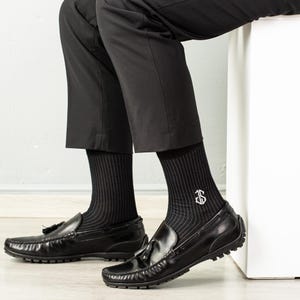 May include: A pair of black dress socks with white pinstripes and a white monogram on one sock. The socks are worn with black loafers.