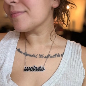 Puede incluir: Un collar de cadena plateada con un colgante de plástico transparente que dice "weirdo" en letras negras. El collar lo lleva una persona con un tatuaje en el pecho que dice "been degraded, We all will be the".