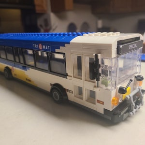 Peut inclure: Un bus jouet blanc, bleu et jaune avec le texte "TRI MET" et "SPECIAL" visible. Le bus a des fenêtres transparentes et des feux orange. C'est un modèle de bus urbain.