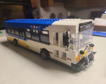 Puede incluir: Un autobús de juguete blanco, azul y amarillo con el texto "TRI MET" y "SPECIAL" visibles. El autobús tiene ventanas transparentes y luces naranjas. Es un modelo de autobús urbano.