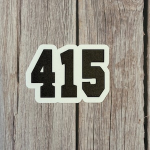 May include: A white sticker with the black numbers "415" in a bold, sans-serif font. The sticker is set against a weathered, light brown wooden background, with visible wood grain and knots.